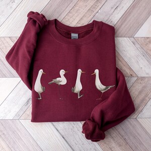 May include: A maroon sweatshirt featuring a watercolor illustration of four stylized birds. The birds are depicted in various poses, with white bodies, gray wings, and yellow beaks. The sweatshirt has a crew neck and long sleeves.