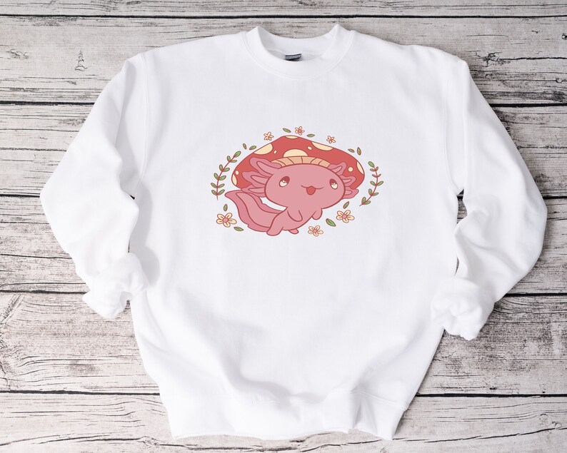 Peut inclure: Sweat-shirt blanc avec un axolotl rose portant un chapeau de champignon rouge et entour&eacute; de fleurs.