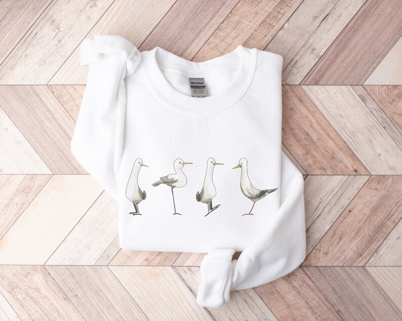 May include: White sweatshirt featuring a watercolor illustration of four seagulls in various poses. The birds have gray wings and are standing on thin black legs. The sweatshirt is made of a soft material and is suitable for casual wear.