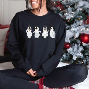 Puede incluir: Una sudadera negra con un gr&aacute;fico de cuatro ping&uuml;inos en fila. Los ping&uuml;inos son blancos con ojos y picos negros. Los ping&uuml;inos llevan bufandas rojas.