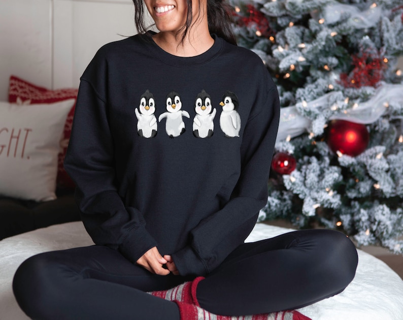 Puede incluir: Una sudadera negra con un gr&aacute;fico de cuatro ping&uuml;inos en fila. Los ping&uuml;inos son blancos con ojos y picos negros. Los ping&uuml;inos llevan bufandas rojas.