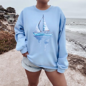 Pode incluir: Uma camisa de frio azul claro com uma pintura de aquarela de um veleiro na frente. O veleiro &eacute; branco com detalhes azuis e se reflete na &aacute;gua.