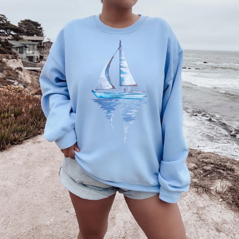 Pode incluir: Uma camisa de frio azul claro com uma pintura de aquarela de um veleiro na frente. O veleiro &eacute; branco com detalhes azuis e se reflete na &aacute;gua.