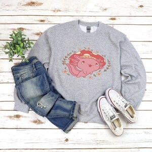 Peut inclure: Un sweat-shirt gris avec un mignon dessin anim&eacute; d'axolotl portant un champignon rouge sur la t&ecirc;te. L'axolotl est entour&eacute; de fleurs et de feuilles. Le sweat-shirt est associ&eacute; &agrave; un jean bleu et des baskets blanches.