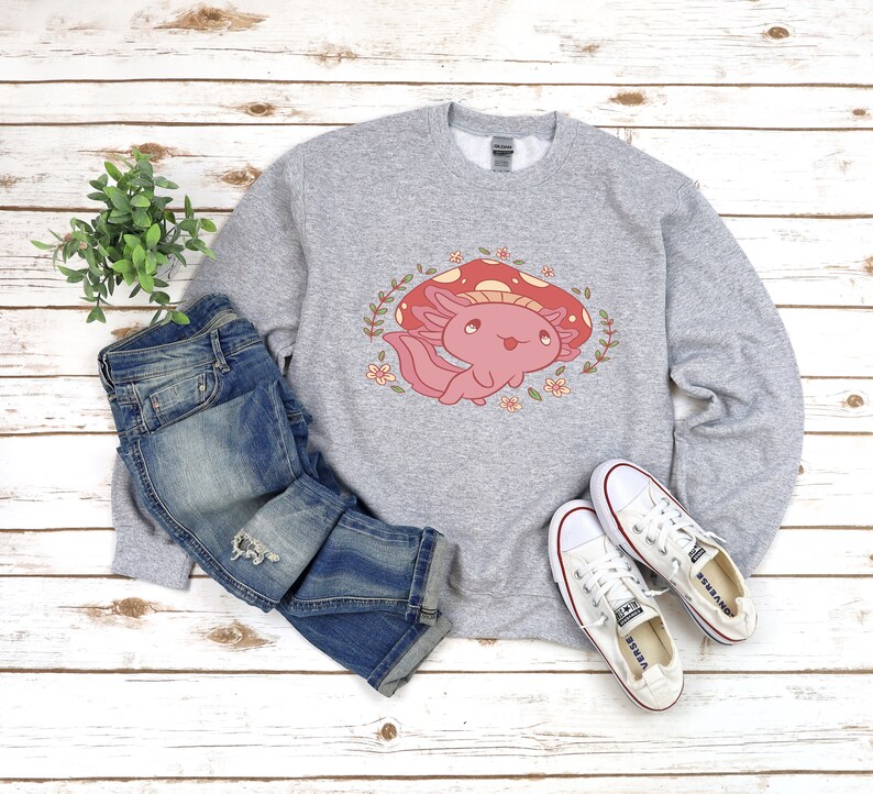 Peut inclure: Un sweat-shirt gris avec un mignon dessin anim&eacute; d'axolotl portant un champignon rouge sur la t&ecirc;te. L'axolotl est entour&eacute; de fleurs et de feuilles. Le sweat-shirt est associ&eacute; &agrave; un jean bleu et des baskets blanches.