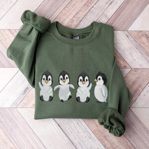 Puede incluir: Una sudadera verde con un gr&aacute;fico de cuatro ping&uuml;inos en fila. Los ping&uuml;inos son blancos con ojos y picos negros, y todos miran hacia adelante. La sudadera tiene cuello redondo y mangas largas.