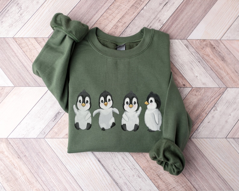 Puede incluir: Una sudadera verde con un gr&aacute;fico de cuatro ping&uuml;inos en fila. Los ping&uuml;inos son blancos con ojos y picos negros, y todos miran hacia adelante. La sudadera tiene cuello redondo y mangas largas.
