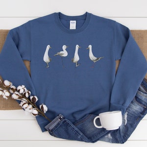 May include: A blue sweatshirt featuring a watercolor design of four seagulls in various poses. The birds are depicted in shades of gray and white. The sweatshirt is displayed with a pair of blue jeans, a white mug, and cotton stems.