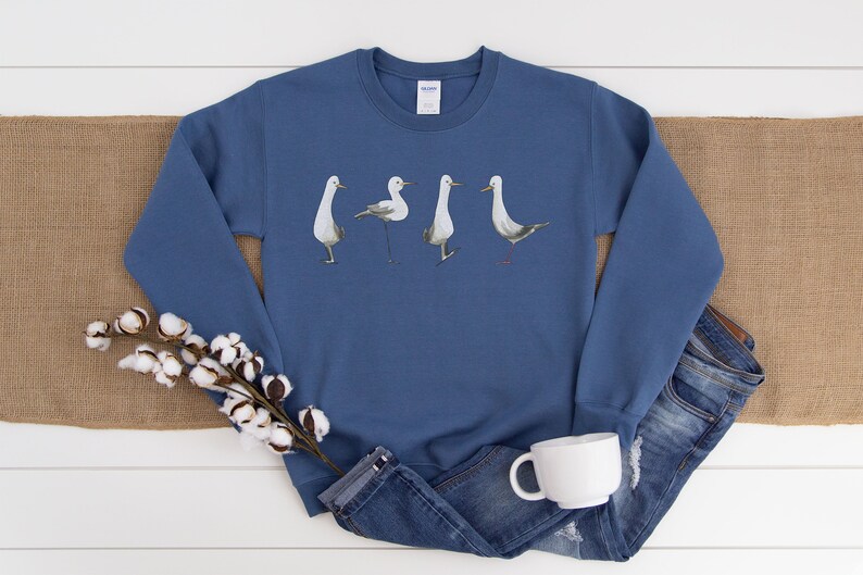 May include: A blue sweatshirt featuring a watercolor design of four seagulls in various poses. The birds are depicted in shades of gray and white. The sweatshirt is displayed with a pair of blue jeans, a white mug, and cotton stems.