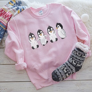 Puede incluir: Una sudadera rosa con un gr&aacute;fico de cuatro ping&uuml;inos caminando en l&iacute;nea. Los ping&uuml;inos son negros y blancos con picos naranjas.