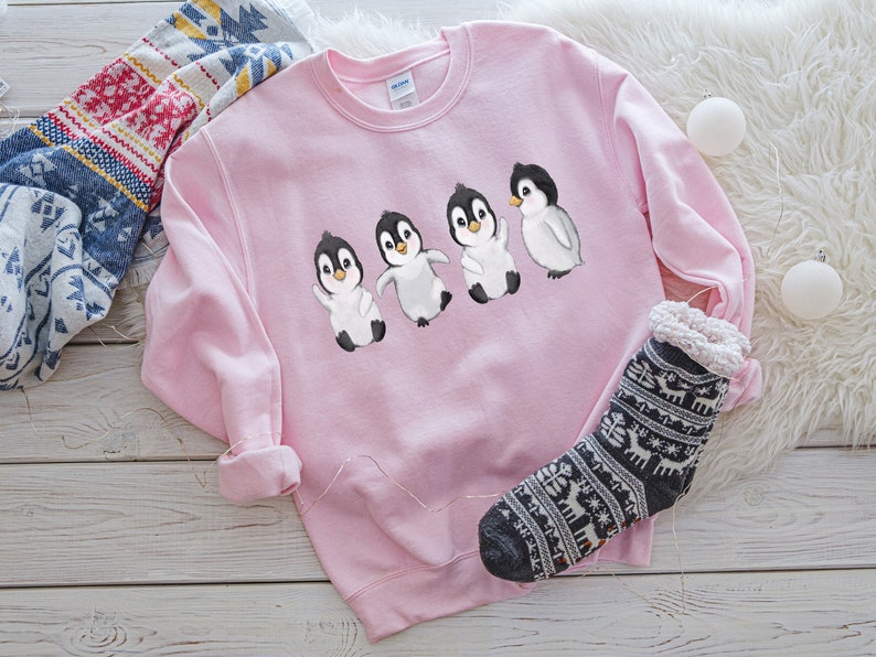 Puede incluir: Una sudadera rosa con un gr&aacute;fico de cuatro ping&uuml;inos caminando en l&iacute;nea. Los ping&uuml;inos son negros y blancos con picos naranjas.