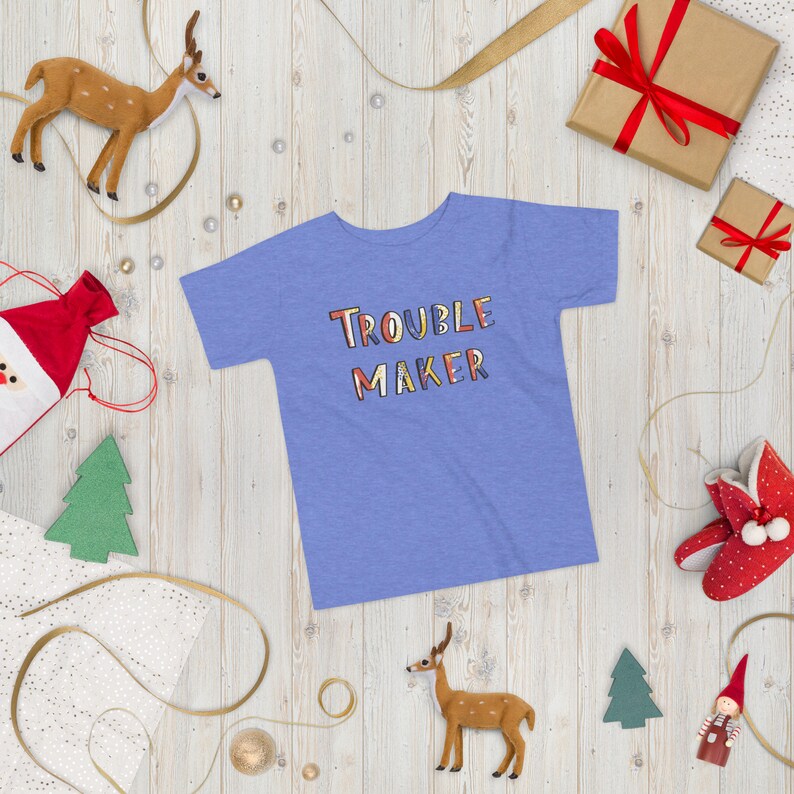 May include: A light blue t-shirt with the words "Trouble Maker" in colorful, stylized lettering. The shirt is on a white wooden background with various decorative items, including a deer figurine, a small Christmas tree, and a gift box.