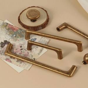 Pode incluir: Conjunto de ferragens para armários em tom bronze, incluindo puxadores e puxadores. O conjunto apresenta puxadores retangulares de vários tamanhos e um puxador redondo, todos com acabamento vintage. Uma fatia de madeira e papel com motivos florais também estão presentes.