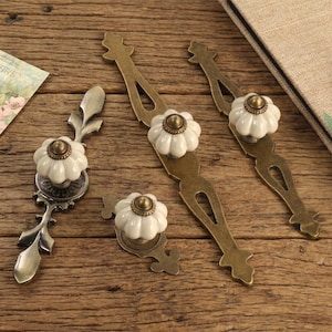 Op de afbeelding: Een verzameling antieke meubelbeslag. Het beslag heeft witte, bloemvormige keramische knoppen met messing accenten en sierlijke metalen achterplaten. Een vintage ansichtkaart met een vogelillustratie en een stoffen boek zijn ook aanwezig.