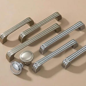 May include: A collection of silver-toned cabinet hardware, including several bar pulls and two round knobs. The pulls have a ribbed design, and the knobs are smooth and circular. The hardware is arranged on a neutral background.