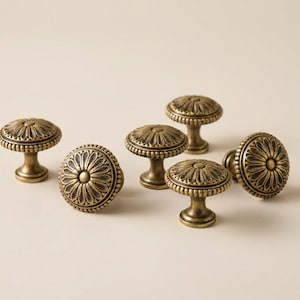 May include: Set of six antique brass cabinet knobs. Each knob features a detailed floral design on the face and a rounded base. The knobs are made of metal and have a warm, aged finish, perfect for adding a vintage touch to furniture.