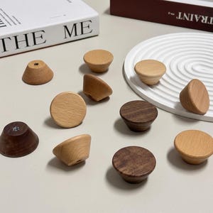 以下が含まれることがあります： さまざまな形と茶色の濃淡の木製キャビネットノブのコレクション。明るい色のノブもあれば、濃い茶色のノブもあります。ノブはニュートラルな表面に配置され、背景には本と装飾トレイがあります。