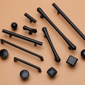 May include: An assortment of black cabinet hardware, including knobs and pulls, in various shapes and sizes. The hardware is displayed on a neutral tan surface, showcasing a modern design aesthetic. The collection includes round, square, and geometric designs.