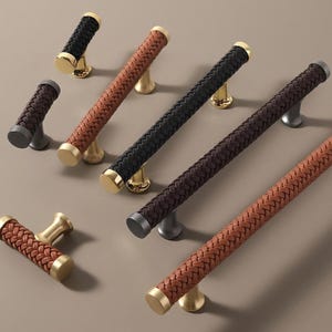 May include: A collection of cabinet pulls and handles in various colors. The handles feature a woven leather design in black, brown, and tan, with gold, silver, and brass-toned bases. The hardware is designed for furniture and home decor.