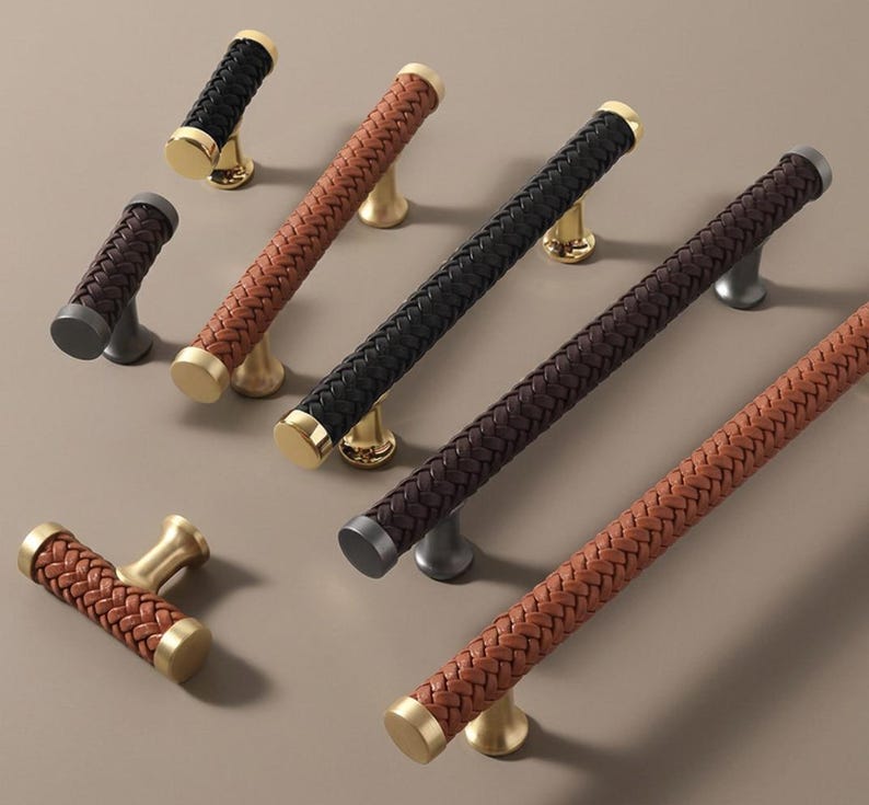 May include: A collection of cabinet pulls and handles in various colors. The handles feature a woven leather design in black, brown, and tan, with gold, silver, and brass-toned bases. The hardware is designed for furniture and home decor.