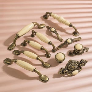 May include: A collection of antique-style cabinet hardware, including handles, knobs, and pulls. The hardware features a cream-colored ceramic or porcelain center with an aged brass finish. Various shapes and sizes are displayed on a pink surface.