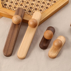 May include: Wooden cabinet pulls in various shades of brown and natural wood. Each pull features a rounded knob. The pulls are arranged on a neutral surface with a decorative wooden frame and a pinecone.