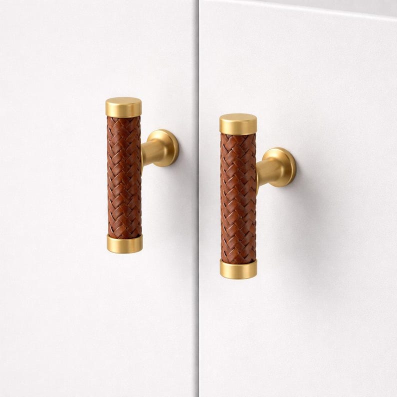 May include: Two cabinet pulls with a woven brown leather center and gold-colored metal accents. The pulls are cylindrical with a T-bar shape, designed for easy gripping. The hardware is set against a white background.