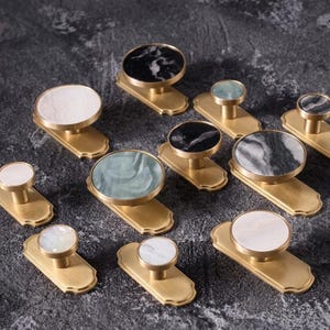 May include: A selection of brass cabinet knobs with decorative tops. The knobs have round tops in various colours and patterns, including white, black, and green marble-like designs. The knobs are mounted on rectangular brass bases.