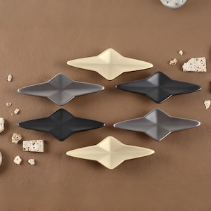 May include: Six star-shaped decorative dishes in shades of black, gray, and cream are arranged on a brown surface. The dishes have a geometric design and are surrounded by small, irregularly shaped stones.