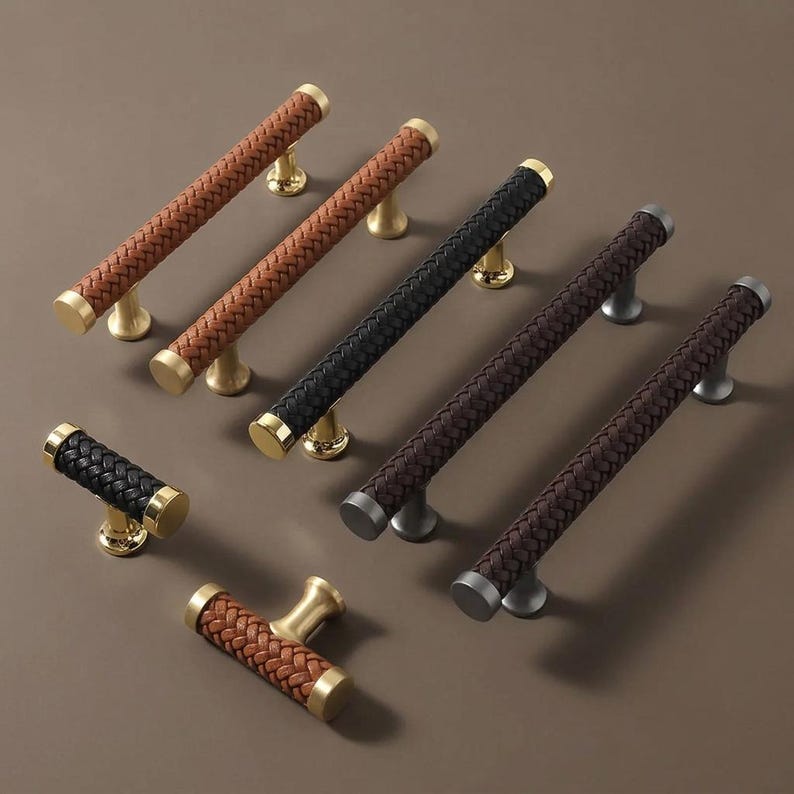 May include: A collection of cabinet pulls with woven leather-like grips in various colors, including brown, black, and dark brown. The pulls have gold or silver-toned bases and end caps, offering a mix of styles for home decor.