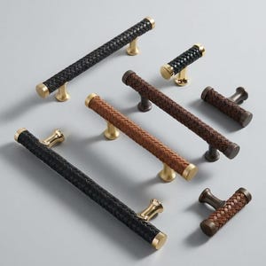 May include: An assortment of cabinet pulls and handles with woven leather-like grips. The hardware features gold and bronze-toned bases and end caps. The collection includes both long bar pulls and smaller knob-style handles in black and brown.