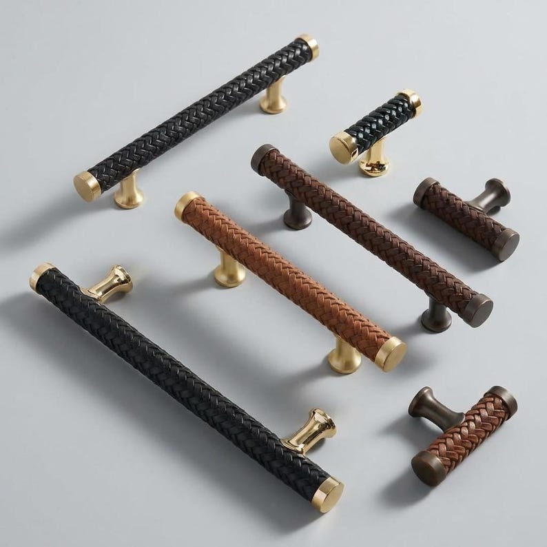 May include: An assortment of cabinet pulls and handles with woven leather-like grips. The hardware features gold and bronze-toned bases and end caps. The collection includes both long bar pulls and smaller knob-style handles in black and brown.