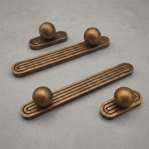May include: Four antique bronze cabinet pulls and knobs. The pulls are rectangular with a grooved design, and the knobs are spherical. The hardware has a warm, aged finish, suggesting a vintage or industrial aesthetic. The pieces are arranged on a neutral gray surface.