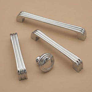 May include: A collection of silver-toned cabinet hardware, including two long handles, one shorter handle, and a round knob. Each piece features a grooved design, reflecting light. The hardware is displayed on a neutral, light brown surface.
