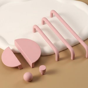 Puede incluir: Un conjunto de herrajes de armario rosas, que incluyen tres tiradores de barra curvados, dos manijas de semicírculo, un pomo redondo y un pomo cilíndrico. Presentado sobre un fondo neutro con una bandeja blanca de forma orgánica.