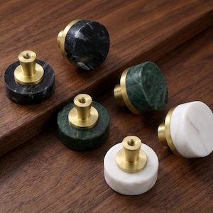 Könnte beinhalten: Eine Auswahl an dekorativen Schrankknöpfen aus Marmor in Schwarz, Grün und Weiß. Jeder Knopf hat einen Messingfuß und -schaft. Die Knöpfe sind auf einer Holzoberfläche angeordnet. Geeignet für Schränke und Schubladen.