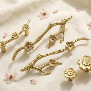 May include: A collection of gold-colored cabinet hardware, including handles and knobs, with a floral and branch design. The hardware is arranged on a cream-colored surface, with scattered flower petals.