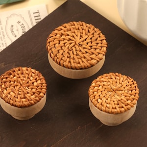May include: Three small, round decorative containers with woven, light brown lids and beige bases. The containers are arranged on a dark brown surface, with a newspaper clipping and a white ceramic pot in the background.