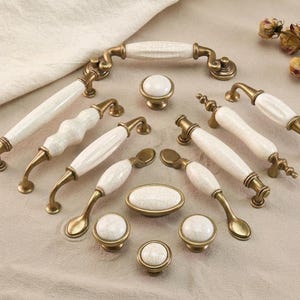 May include: A collection of antique-style cabinet hardware, including handles and knobs. The hardware features white ceramic or porcelain accents with a gold-toned metal finish. Various shapes and sizes are displayed, suitable for furniture or cabinetry.