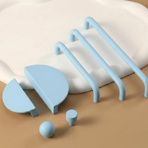 May include: A selection of light blue cabinet hardware, including three curved bar pulls, two half-circle pulls, a round knob, and a small cylindrical knob. The hardware is displayed on a neutral surface with a white, organic-shaped tray in the background.