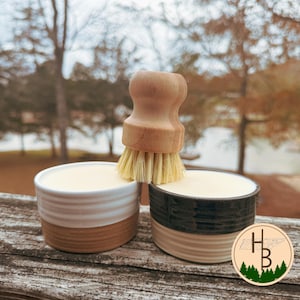 Può includere: Una spazzola per piatti con manico in legno e setole colorate chiare poggia su due ciotole di ceramica piene di una sostanza cremosa. Una ciotola è bianca e marrone, l'altra è nera e beige. Lo sfondo è sfocato con alberi e acqua.
