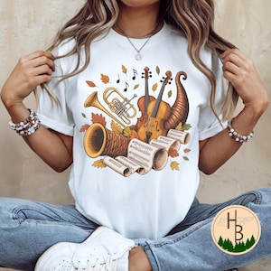 May include: White t-shirt with a musical design. It features a trumpet, violin, and musical notes, alongside autumn leaves and a cornucopia. The artwork uses warm colours, creating a fall or harvest aesthetic.