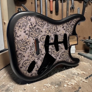 Könnte beinhalten: Ein E-Gitarrenkorpus mit mattschwarzen Kanten und einer hellbeigen Vorderseite, die mit einem dunkelbraunen und schwarzen Paisley-Muster verziert ist. Der Korpus verfügt über Aussparungen für Tonabnehmer, Steg und Halsbefestigung.