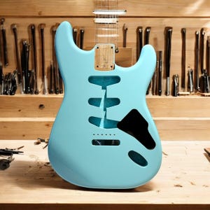 Könnte beinhalten: Ein hellblauer E-Gitarrenkorpus mit schwarzem Schlagbrett und einem Holz Hals. Die Gitarre liegt auf einer Holzwerkbank.