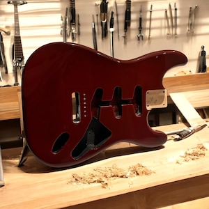 Könnte beinhalten: Ein glänzender, burgunderfarbener Korpus für eine E-Gitarre mit Aussparungen für Tonabnehmer und Bedienelemente. Der Korpus ist unfertig und liegt auf einer Holzwerkbank. Werkzeuge sind im Hintergrund zu sehen.