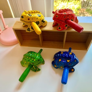 May include: Four wooden frog shakers, each painted in a different color: yellow, red, green, and blue. The frogs have black spots and are sitting on a wooden tray.