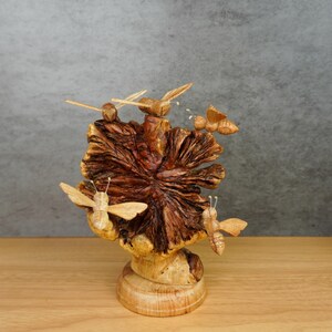 May include: A hand-carved wooden sculpture featuring a central dark brown textured burl wood piece shaped like a flower or fan, mounted on a light-colored circular wooden base, with four small carved wooden bees attached to the structure.