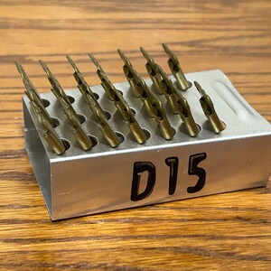May include: A metal holder with multiple drill bits. The drill bits are gold-colored and arranged in a grid. The holder is silver and has the number "D15" printed on the side. The background is a wooden surface.