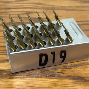 May include: A metal container holding multiple gold-colored drill bits. The container is silver and has the number "D19" printed on the side. The drill bits are arranged in rows and are angled upwards.