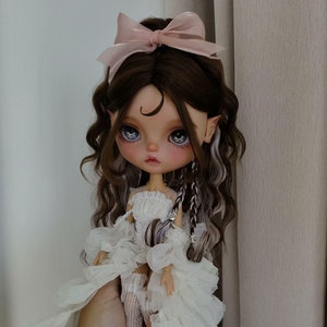 May include: A white doll with long brown and white hair, wearing a white dress with ruffles and a pink bow. The doll has blue eyes and a painted crescent moon on her forehead.
